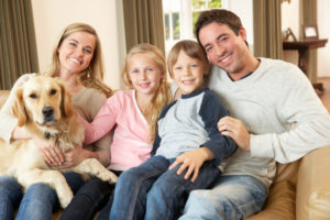 Happy Young Family Sitting On Sofa Holding A Dog